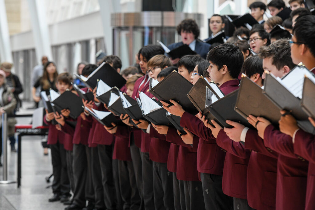 A group of young male students in maroon and black uniforms standing in rows inside a modern building, holding black choir folders and singing. The focus is on the students at the front, with blurred onlookers in the background.