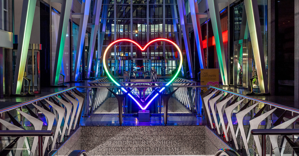A vibrant entrance to Allen Lambert Galleria at night, showcasing a large neon-lit heart in rainbow colors, flanked by sleek, metallic pillars and illuminated architectural details. Below the heart, a sign reads 'Food Court & Shops, Hockey Hall of Fame,' emphasizing the location's attractions.