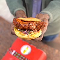 A person holding a crispy chicken sandwich with a rich, dark crust and fresh vegetables on a bun, over a blurred background featuring a red food payment box.