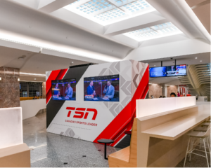 A sleek and contemporary sports media space within an indoor setting, featuring a large, angular media wall branded with 'TSN - Canada's Sports Leader.' The wall displays multiple screens showing sports analysts in discussion, enhancing the dynamic atmosphere.