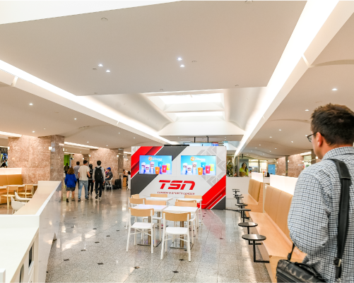 A vibrant, open sports media area inside a modern shopping center, featuring a prominent red and white media wall with the TSN logo and multiple screens broadcasting live sports. The area is equipped with minimalist white tables and seating, providing a communal spot for shoppers to catch up on sports. The busy corridor surrounding the media area adds a dynamic element, with visitors walking by, some glancing towards the action on the screens.