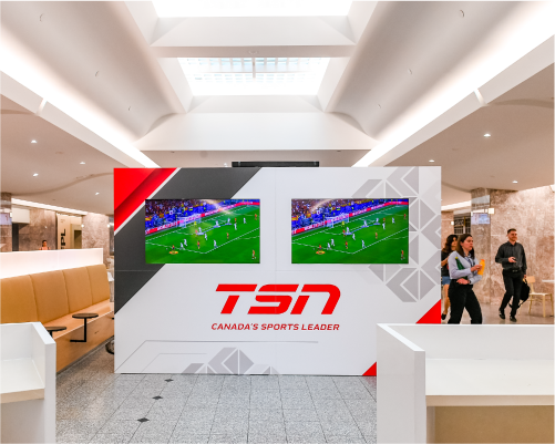 A detailed view of a red and white sports media wall branded with 'TSN - Canada's Sports Leader' and showcasing two large screens broadcasting soccer matches. This setup forms a focal point in a public area, surrounded by simple yet stylish seating arrangements that invite visitors to watch and enjoy the games.