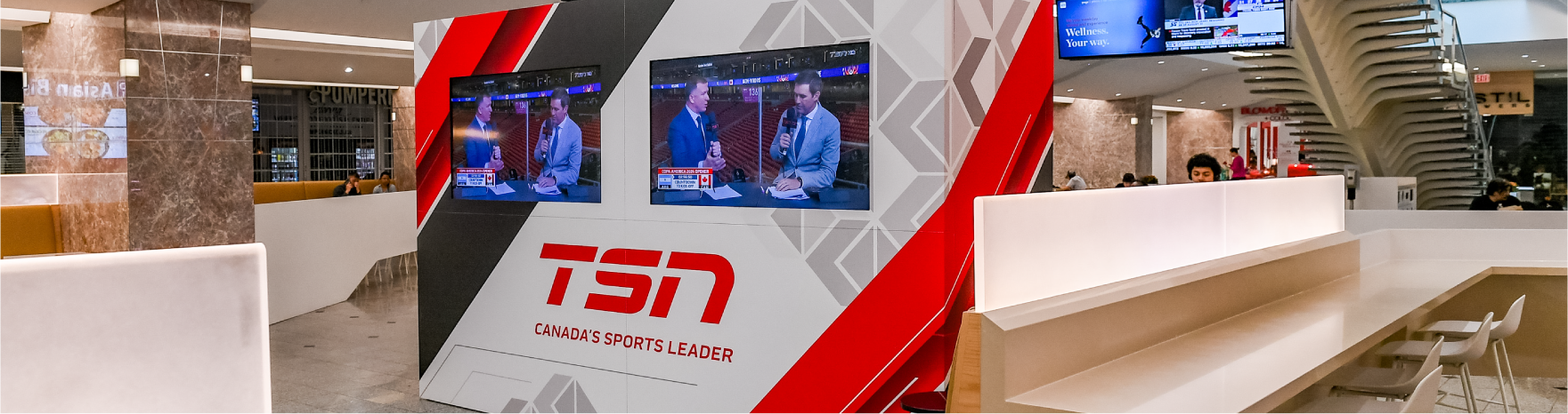 A sleek and contemporary sports media space within an indoor setting, featuring a large, angular media wall branded with 'TSN - Canada's Sports Leader.' The wall displays multiple screens showing sports analysts in discussion, enhancing the dynamic atmosphere.