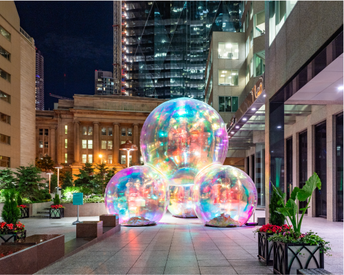 An enchanting view of a public art installation at night, featuring three large, interconnected orbs that radiate a spectrum of iridescent colors. Situated in an urban plaza, the orbs are beautifully highlighted against the contrasting architecture of modern glass buildings and a historic stone structure. Lush greenery and vibrant red flowers in planters add a touch of natural beauty to the scene, creating a magical urban oasis under the city lights.