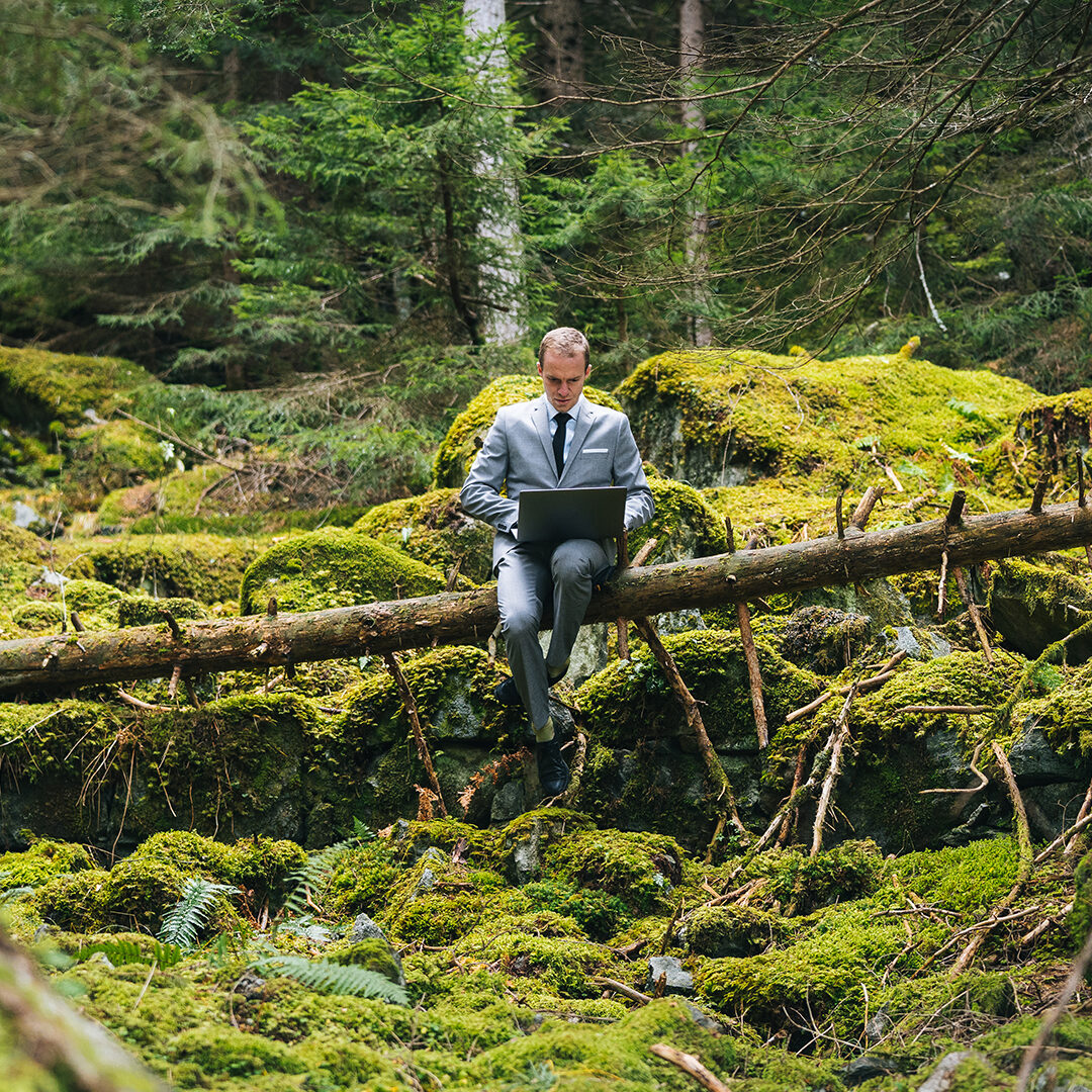 Decorative image of a business man in a grey suit with black tie, sitting on a fallen tree in the forest doing work on his laptop