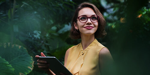 Decorative image of a woman with short brown hair and glasses folder a clipboard while standing in a greenhouse.