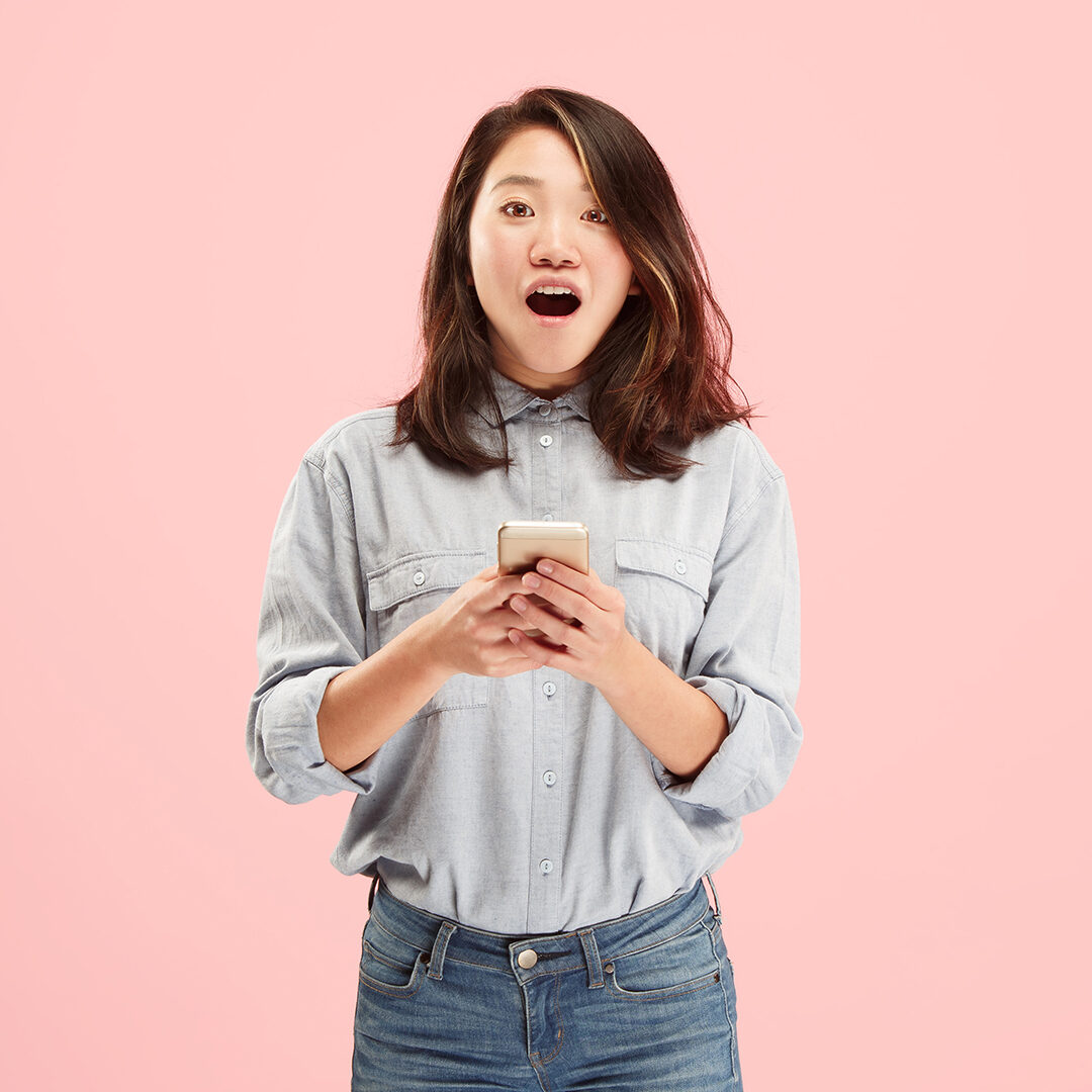 Decorative image of a happy young woman in a canadian tuxedo holding a phone. she seems very excited by what is on the phone. The background of the image is a solid pale salmon colour.