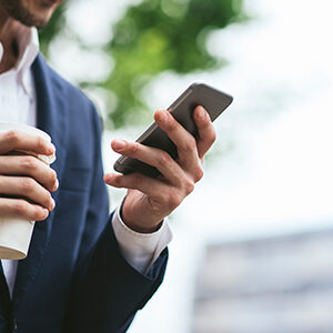 Close croped photograph of a business man's hands and torso. He is wearing a suit and holding both a coffee cup and a smart phone